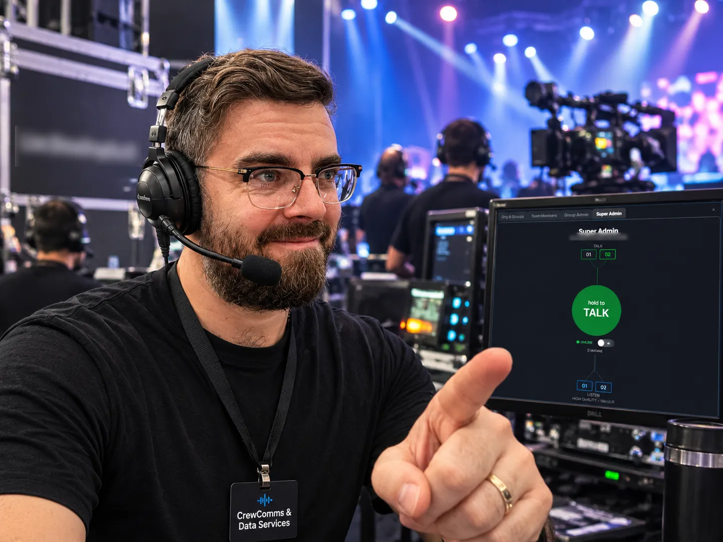 A crew member in a headset at a live event control desk, pointing to the comms app on a monitor